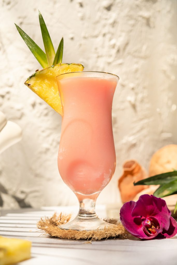 hand grabbing pink Aperol Piña Colada in a hurricane cocktail glass with aperol bottle and tropical fruit