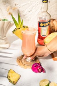 hand grabbing pink Aperol Piña Colada in a hurricane cocktail glass with aperol bottle and tropical fruit