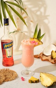 pink Aperol Piña Colada in a hurricane cocktail glass with aperol bottle and tropical fruit